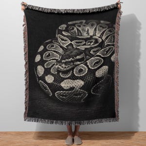 Op de afbeelding: Zwart-wit sprei met een opgerolde slang met een gedetailleerd patroon van vlekken en schubben. De kop van de slang is zichtbaar, met de ogen naar voren gericht. De deken heeft een gefranjerde rand.