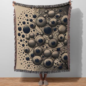 May include: A beige throw blanket with a repeating pattern of dark blue and gray spheres connected by lines. The blanket has a fringe border in shades of brown and black. The design is reminiscent of a scientific illustration.