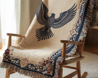 May include: A woven throw blanket featuring a large, dark blue eagle with wings spread wide against a light beige background. The blanket has a decorative dark blue border with floral patterns and fringe along the edges, draped over a wooden chair.