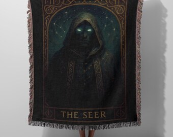 May include: A black woven throw blanket featuring a hooded figure with glowing eyes centered within an arched frame. The design includes celestial star patterns and decorative border motifs. The text THE SEER is printed in gold at the bottom.