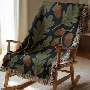 May include: A black throw blanket draped over a wooden rocking chair. The blanket features a repeating pattern of green oak leaves and brown acorns. The blanket has a fringed edge with multiple colors.