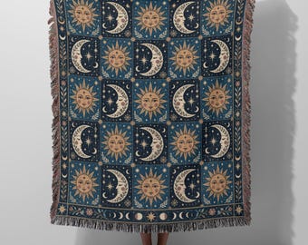 May include: A woven throw blanket featuring a repeating grid pattern of suns and crescent moons in shades of navy blue, cream, and gold. The design includes star accents and floral motifs, finished with a decorative fringed edge on all sides.