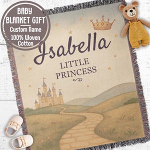 Può includere: Una coperta per bambini tessuta a tema fiabesco. La coperta presenta un castello, un sentiero tortuoso e il nome "Isabella" con "Little Princess" scritto sotto. Un orsacchiotto e scarpine sono accanto.