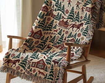 May include: A woven throw blanket featuring a winter scene with dark green pine trees, red houses, a horse-drawn sleigh, ice skaters, and wildlife on a cream background. The blanket has fringed edges and is draped over a wooden rocking chair.