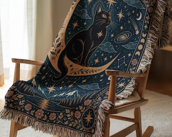 May include: A woven throw blanket featuring a dark blue background with a central black cat sitting on a crescent moon. The design includes stars, a galaxy, clouds, mountains, and floral borders with fringed edges on the sides and bottom.