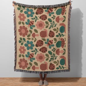 Puede incluir: Manta beige con un patrón repetido de flores, mariquitas, abejas, caracoles y setas en tonos marrón, rosa, verde azulado y verde. La manta tiene un borde con flecos y la sostiene una persona.