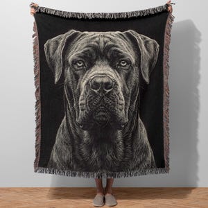 Puede incluir: Manta tejida negra con una ilustración detallada de la cara de un perro. El perro tiene una expresión seria, con pelaje y rasgos faciales detallados. La manta tiene un borde con flecos en tonos marrones y negros, y la sostienen dos personas.