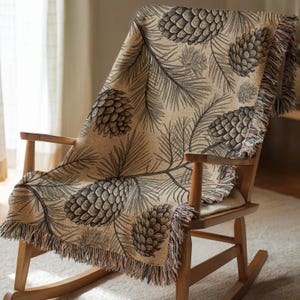 May include: A beige throw blanket with a repeating pattern of pine cones and pine needles draped over a wooden rocking chair. The blanket has a fringed edge with multiple colors. The chair is made of light-colored wood.
