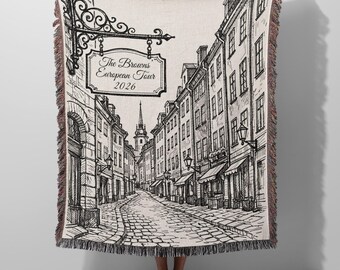 May include: A woven throw blanket featuring a black and white line art illustration of a European street scene with cobblestone paths and buildings. A hanging sign reads The Browns European Tour 2026. The blanket has a fringed edge on all sides.