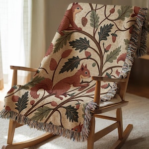 Peut inclure: Plaid beige avec un motif répété d'écureuils bruns, de glands et de feuilles. La couverture est drapée sur un fauteuil à bascule en bois. La couverture a une bordure à franges.