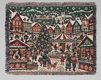 May include: A woven throw blanket featuring a detailed winter scene of a Christmas market with a carousel, a large decorated tree, snow-covered houses, and people walking through the village. The design includes red, green, and white colours with fringed edges.