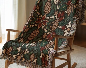 May include: A woven throw blanket featuring a dark background with a repeating pattern of pinecones, evergreen branches, red berries, and white mistletoe. The blanket has a fringed edge and is draped over a wooden rocking chair.