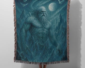 May include: A rectangular woven throw blanket featuring a dark blue, monochromatic illustration of a bearded, muscular giant standing amidst jagged mountain peaks under a full moon, with two flying birds and decorative fringe along the edges.