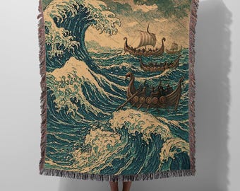 May include: A woven throw blanket featuring a detailed illustration of multiple Viking longships navigating through large, turbulent ocean waves. The design uses a palette of deep teal, dark blue, and cream, finished with a decorative fringed edge.