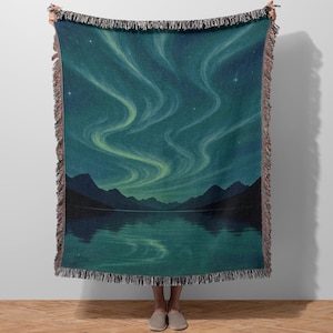 May include: A woven throw blanket featuring a scenic illustration of the Northern Lights over a lake and mountains. The design showcases swirling green and white lights against a dark blue sky, with a mountain range reflected in the water. The blanket has a fringed edge.
