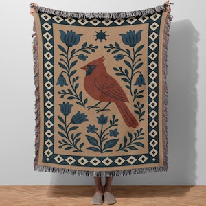 May include: A woven throw blanket featuring a cardinal bird design. The blanket has a tan background with a border of blue and white diamonds. The cardinal is brown with a black head, surrounded by blue floral elements.