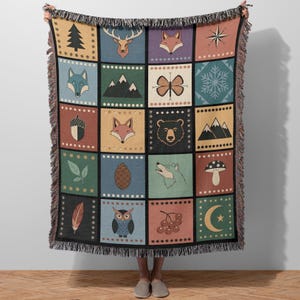May include: A woven throw blanket with a patchwork design featuring forest-themed illustrations. The squares display images of a deer, fox, bear, mountains, and other nature elements. The blanket has a fringed edge and is held up by a person.