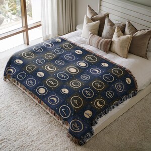 May include: A navy blue throw blanket with a repeating pattern of white celestial symbols and runes. The blanket has a fringed edge and is displayed on a bed with pillows. The design includes moons, stars, and other mystical elements.