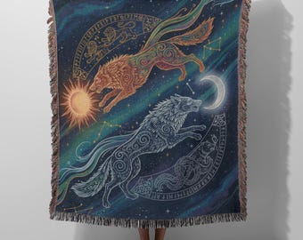 May include: A woven throw blanket featuring a dark blue celestial background with two wolves, one orange and one blue, leaping across the fabric. The design includes a sun, a crescent moon, star constellations, and circular borders with runic symbols.