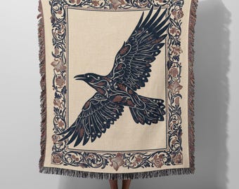 May include: A woven throw blanket featuring a central dark blue raven in flight with intricate leaf patterns on its wings. The blanket has a cream-colored background and a decorative border of dark blue and brown floral motifs with fringed edges.