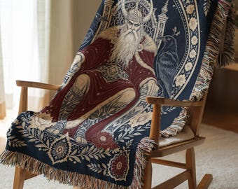 May include: A woven throw blanket featuring a central illustration of a bearded figure in a horned helmet sitting on a throne, flanked by two ravens. The design includes deep red, navy blue, and cream floral borders with decorative fringe edges.