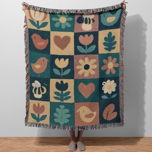 May include: A woven throw blanket with a patchwork design featuring birds, flowers, hearts, and bees in a grid pattern. The colour palette includes teal, tan, and muted pink. The blanket has a fringed edge.