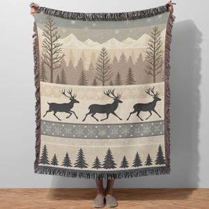 Könnte beinhalten: Eine gewebte Wurkdecke mit einer Winterwaldszene. Das Design zeigt eine Bergkette, Bäume, Schneeflocken und Silhouetten von laufenden Hirschen in Grau-, Beige- und Brauntönen. Die Decke hat einen Fransenrand.