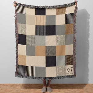 May include: A patchwork throw blanket featuring geometric patterns in black, grey, beige, and cream. The blanket has a fringed edge and the initials "K & T" with "EST. 2023" are printed on the lower right corner. The blanket is held up by a person.