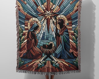 Op de afbeelding: Een geweven sprei met een afbeelding van de Geboorte van Christus in glas-in-loodstijl. Het ontwerp toont Maria, Jozef en het kindje Jezus, met een ster en geometrische vormen in blauw, bruin en goud. De sprei heeft een gefranjerde rand.