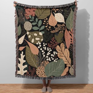 May include: A black throw blanket with a botanical pattern featuring various leaves and floral elements in shades of green, brown, and beige. The blanket has a fringed edge and is held up by a person.