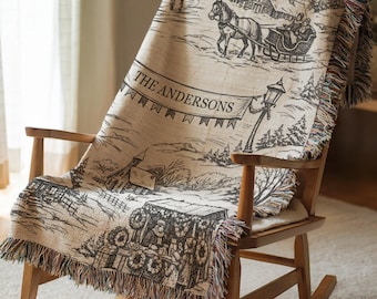 May include: A woven throw blanket featuring a winter scene with a horse-drawn sleigh, a street lamp, and a market stall. The text THE ANDERSONS is printed on a banner across the center. The blanket has fringed edges and a beige and black color scheme.
