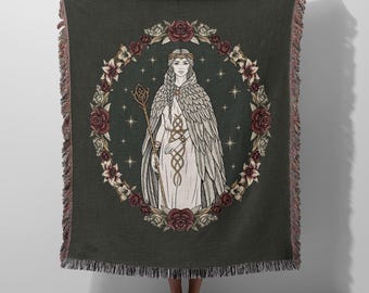 May include: A dark gray woven throw blanket featuring a central illustration of a figure in a long white gown and feathered cloak holding a staff. The figure is encircled by a wreath of red roses and white lilies against a backdrop of stars.
