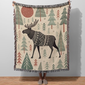 May include: Beige woven throw blanket with a forest and moose design. The blanket features a black moose wearing a patterned sweater, surrounded by green and red trees, and a red sun. The edges have a black fringe.
