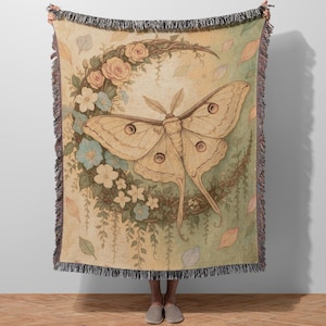 May include: A beige throw blanket featuring a large moth illustration with a floral crescent design. The moth has detailed wings with eye-like markings. The blanket has a fringe border and a soft, vintage aesthetic.
