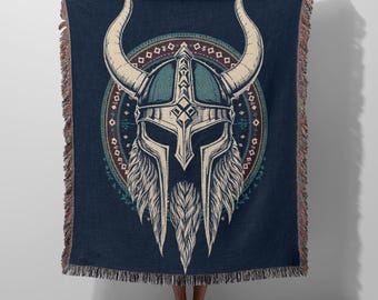 May include: A dark blue woven throw blanket featuring a large, central graphic of a horned Viking helmet with a detailed beard. The design is framed by a circular, patterned border in shades of teal, brown, and white, with fringed edges on all sides.