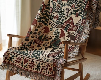 May include: A woven throw blanket featuring a winter scene with gnomes, a moose, and a snowy village. The design includes red, green, and cream colours with a fringed edge. The pattern depicts gnomes carrying lanterns and bowls, a rabbit, and hearts.