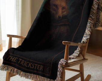 May include: A woven throw blanket featuring a dark, moody illustration of a fox with orange fur and a black cloak. The text THE TRICKSTER is printed in a serif font at the bottom. The blanket has a black background and multi-colored fringe edges.