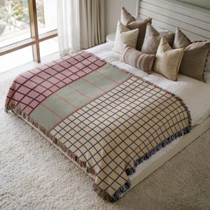 May include: A woven throw blanket with a geometric pattern in shades of pink, green, and beige. The blanket is rectangular with a grid design and fringed edges. It is displayed on a bed with pillows.