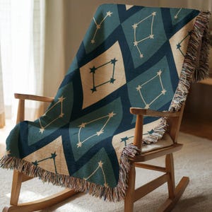 Peut inclure: Une couverture tissée orne un fauteuil à bascule en bois. Le motif géométrique de la couverture présente des losanges et des constellations d'étoiles en bleu canard, beige et bleu marine. Les bords sont ornés de franges.