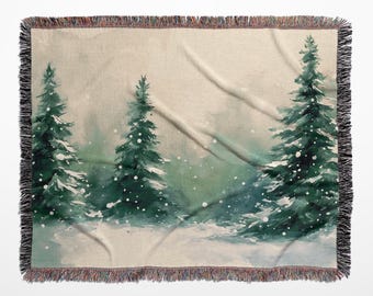 May include: A woven throw blanket featuring a watercolour-style winter forest scene with three dark green pine trees set against a light beige background. The design includes white flecks representing falling snow and a decorative fringed edge.