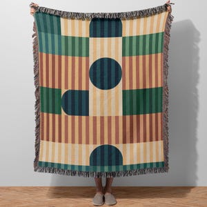 May include: A woven throw blanket with a geometric pattern. The design features vertical stripes in shades of brown, yellow, green, and teal, interspersed with geometric shapes. The blanket has a fringed edge and is held up by a person.