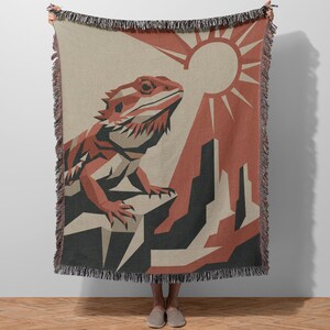 May include: A woven throw blanket with a geometric design of a bearded dragon, sun, and desert landscape in red, black, and beige. The blanket has a fringed edge.