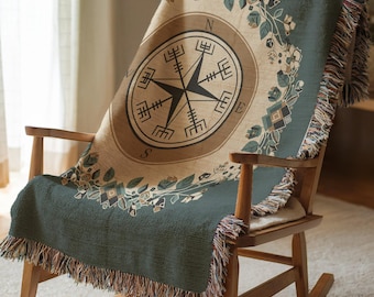May include: A woven throw blanket featuring a central compass design with the letters N, S, E, and W. The blanket has a tan center, a dark green border, and a decorative floral wreath pattern. It is draped over a wooden rocking chair with fringe edges.