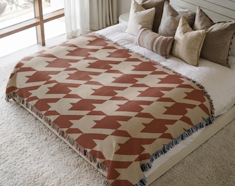 May include: A woven throw blanket featuring a repeating geometric houndstooth pattern in terracotta orange and cream. The rectangular blanket has a fringed edge with visible dark blue and cream threads along the bottom border.