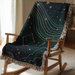 Op de afbeelding: Een donkerblauwe sprei met een hemels ontwerp met sterrenbeelden, sterren en gebogen lijnen in goud- en groentinten. De sprei heeft een gefranjerde rand en is gedrapeerd over een houten schommelstoel.