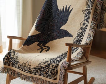 May include: A woven throw blanket featuring a large, dark, stylized raven in flight against a light beige background. The blanket is bordered by a dark, intricate floral pattern and finished with a fringed edge along the bottom and sides.