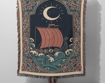 May include: A woven throw blanket featuring a dark blue background with a central illustration of a Viking ship sailing on stylized teal waves under a crescent moon. The design is framed by a decorative border of intricate knots and floral motifs.