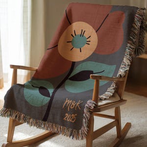 Op de afbeelding: Een decoratieve sprei met een bloemenpatroon, gedrapeerd over een houten schommelstoel. De sprei heeft een groot bloemmotief in bruin-, teal- en oranjetinten op een donkergrijze achtergrond. De tekst "M&K 2025" is ook aanwezig.
