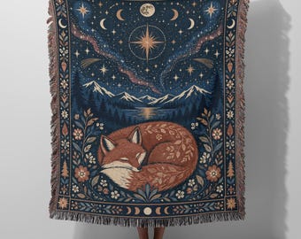 May include: A woven throw blanket featuring a sleeping fox curled in the centre, surrounded by floral patterns. The background depicts a dark blue night sky with a large star, moon phases, and mountains above a forest. It has a fringed edge.