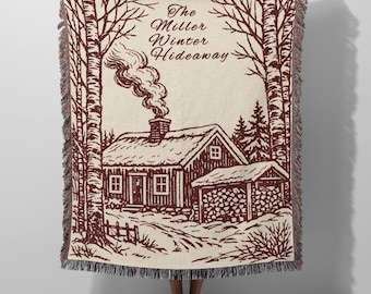 May include: A cream-colored woven throw blanket featuring a dark red illustration of a winter cabin scene with trees, a smoking chimney, and a wood shed. The text "The Miller Winter Hideaway" is printed at the top. The blanket has fringed edges.
